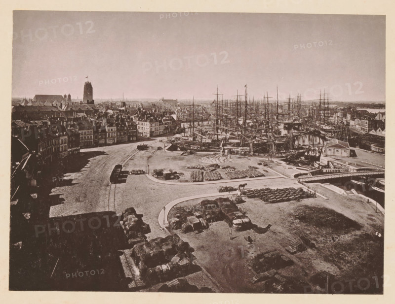 Dunkerque, Vue Générale, View of Dunkirk, page, photomechanical print, inscription: 'Le Nord Contemporain, Revue Pittoresque', photographer: NC, Dunkirk, c. 1878 - in or before 1883-02-20, paper, collotype, photoprint, height 165 mm x width 219 mm, Print on fifth unnumbered page after page 48., prospect of city, town panorama, silhouette of city, A wide waterfront scene shows a harbor filled with tall ships with masts and rigging clustered along the quay, adjacent to rows of multi-story buildings with pitched roofs and chimneys; an open cobblestone square in the foreground contains stacked crates and covered bundles, horse-drawn carts, and a curved roadway leading toward the docks, while a bridge spans a waterway near a small building at the edge of the quay and a distant tower rises above the town skyline.