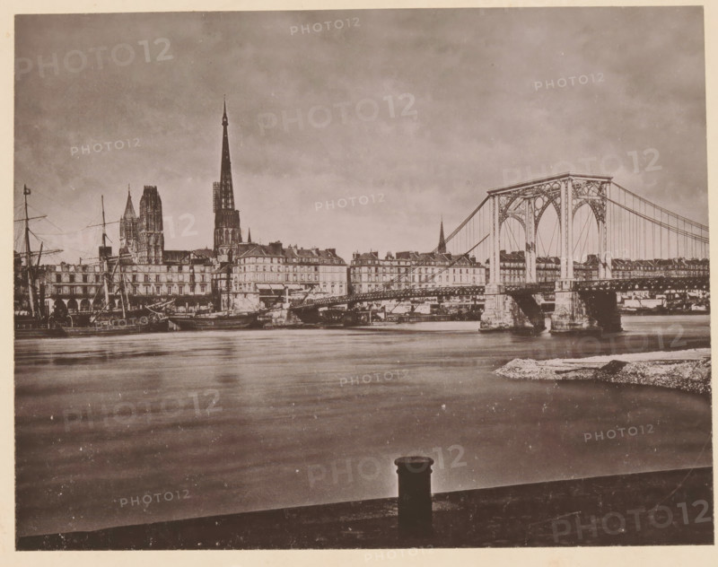 Rouen, Le Pont Suspendu, View of the suspension bridge in Rouen, page, photomechanical print, inscription: 'Le Nord Contemporain, Revue Pittoresque', inscription: 'Journal le Nord Contemporain, No 30, année 1883.', photographer: NC, c. 1878 - in or before 1883-04-05, paper, collotype, photoprint, height 169 mm x width 219 mm, Print on fifth unnumbered page after page 96., prospect of city, town panorama, silhouette of city, suspension bridge, A sepia-toned riverside scene showing calm water in the foreground and a city skyline along the far bank, with a large iron suspension bridge spanning from right to center, ornate towers and cables visible, multi-story buildings lining the riverbank behind the bridge, tall cathedral spires rising above the rooftops toward the left, masts and rigging of docked ships near the left edge, stone bridge piers and quay structures at the waterline, and a bollard silhouetted on the near edge of the riverfront.