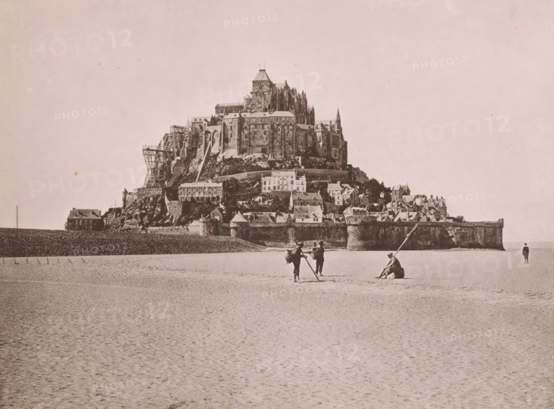 View of Mont Saint-Michel, Le Mont Saint-Michel, Au Péril de la Mer, page, photomechanical print, inscription: 'Le Nord Contemporain, Revue Archéologique & Pittoresque', inscription: 'Journal le Nord Contemporain, No 31, année 1883.', photographer: NC, Mont-Saint-Michel (abbey), c. 1878 - in or before 1883-04-20, paper, collotype, photoprint, height 165 mm x width 219 mm, Print on fifth unnumbered page after page 112., island, prospect of village, silhouette of village, A large rocky island topped by a complex stone building with towers, spires, and scaffolding rises from a flat sandy foreground, with terraces of smaller buildings and defensive walls along its base; in the foreground on the sand three people stand and one sits near long poles while another person stands alone near the waterline, and a solitary house sits at the far left against a low seawall.