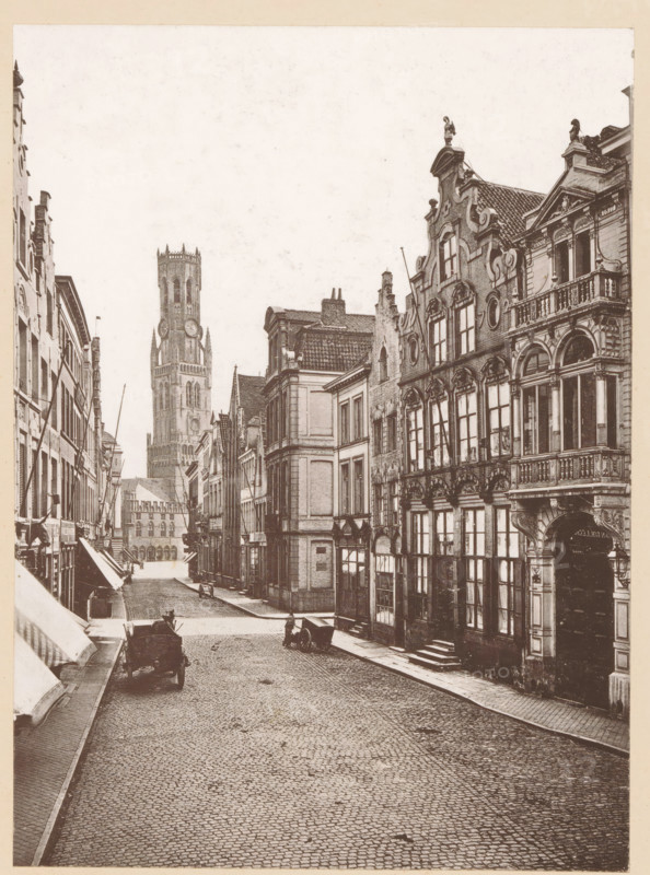 Bruges, La Rue Flamande, View of the Rue Flamande in Bruges, page, photomechanical print, inscription: 'Le Nord Contemporain, Revue Artistique & Archéologique', inscription: 'Journal le Nord Contemporain, No 32, année 1883.', photographer: NC, c. 1878 - in or before 1883-05-05, paper, collotype, photoprint, height 222 mm x width 162 mm, Print on third unnumbered page after page 128., street, A narrow cobbled street lined with tall, ornate buildings on both sides, featuring decorative facades, arched windows, balconies, and sculpted rooflines; a tall tower with a clock face rises in the distance, flanked by other stone structures; a few horse-drawn carts and a person near one cart are visible on the street, and awnings extend from some shopfronts along the left side.