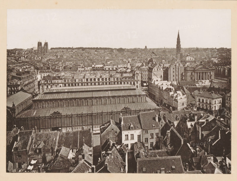 View of Brussels, Bruxelles, Vue Générale, page, photomechanical print, inscription: 'Journal le Nord Contemporain, No 34, année 1883.', inscription: 'Le Nord Contemporain, Revue Artistique & Archéologique', photographer: NC, Brussels, c. 1878 - in or before 1883-06-05, paper, collotype, photoprint, height 160 mm x width 220 mm, Print on third unnumbered page after page 160., prospect of city, town panorama, silhouette of city, A wide aerial view of an urban area showing many tightly packed buildings with varied rooflines and tiled roofs in the foreground, a large long building with a metal or glass roof and a colonnaded facade occupying the midground, rows of multi-story masonry buildings with ornate façades and dormer windows behind it, and a distant cityscape with taller church spires and towers rising above the lower rooftops under a pale sky.
