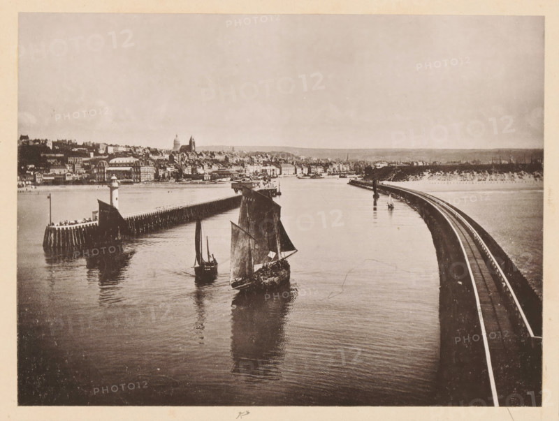 View of Boulogne-sur-Mer, Boulogne-sur-Mer, Vue Générale, page, photomechanical print, inscription: 'Le Nord Contemporain, Revue Artistique & Archéologique', inscription: 'Journal le Nord Contemporain, No 34, année 1883.', photographer: NC, c. 1878 - in or before 1883-06-05, paper, collotype, photoprint, height 164 mm x width 218 mm, Print on fifth unnumbered page after page 160., prospect of city, town panorama, silhouette of city, coastal structures, A sepia-toned waterfront scene shows calm water reflecting boats with tall sails in the foreground, a curved railway embankment running along the right side toward the distance, and a timber breakwater with a small lighthouse structure on the left. Along the far shore a dense cluster of buildings rises behind the waterfront, including larger domed and towered structures, with a low horizon and open sky above.
