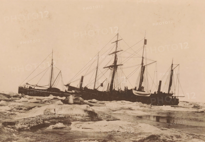 The Dymphna and the Varna shortly after freezing. September 23 1882., View of the Dymphna and the Varna in the Kara Sea, page, photomechanical print, inscription: 'De Nederlandsche Polar-Expeditie 1882-1883.', photographer: H. Ekama, Karskoye More, cliché maker: E. Diener, Glauchau, c. 1881 - in or before 1886, Saxony, paper, collotype, photoprint, height 140 mm x width 206 mm, ships (in general), landscapes in polar regions, A sepia-toned scene showing several docked sailing ships viewed from the side, with tall masts, rigging, and spars silhouetted against a pale sky, small lifeboats hanging from davits on one vessel, decks with indistinct figures and equipment, a rocky foreground with ice floes and textured water, and decorative border details along the top edge of the image.