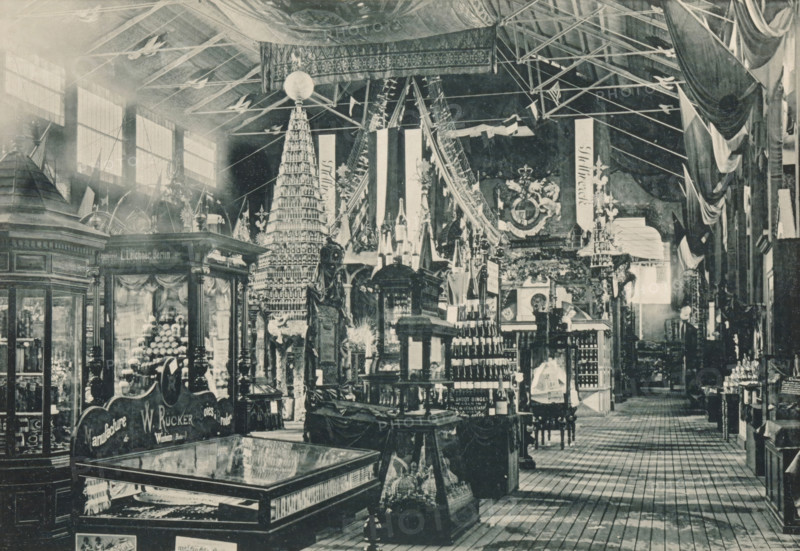 Coup d'oeil sur la section Allemande, Galerie Principale, View of the German part of the main exhibition of the Antwerp World Exhibition in 1894, Exposition Universelle d'Anvers 1894, page, photomechanical print, photographer: Charles Bernhoeft, Antwerp, Luxembourg (Belgian province), 1894, paper, collotype, photoprint, height 152 mm x width 222 mm, counter, shop, world fair, world exhibition, World Exhibition 1894 (Antwerp), Period, 1894-05-05 - 1894-11-05, A large indoor exhibition space filled with ornate wooden display cases, shelves, and decorative booths arranged on a patterned wooden floor. Tall vertical banners hang from the ceiling alongside draped fabric and structural beams, and a high roof with visible rafters and large windows lets light into the hall. Central displays include a tall conical tower made of stacked items topped with a spherical object, rows of bottles and containers on shelves, sculptural figures and carved pedestals, and glass-topped cabinets showing items ...