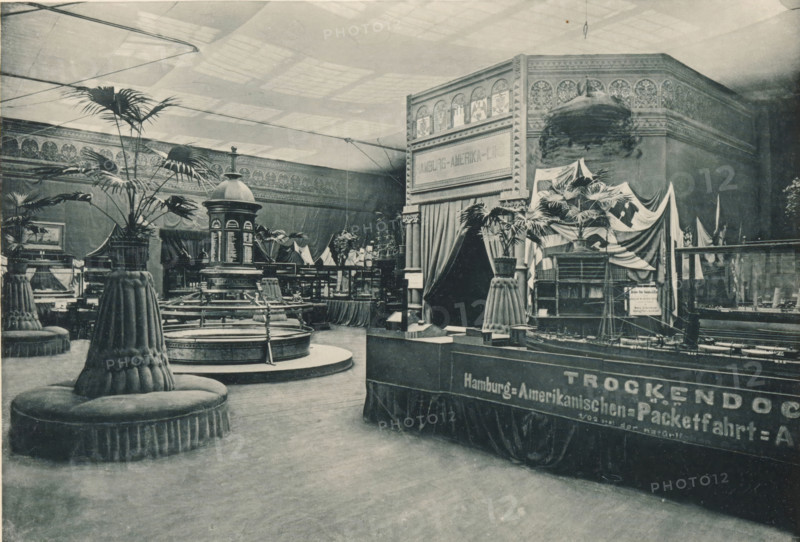 View of the maritime Hamburg part of the Antwerp World Exhibition in 1894, Section Maritime Hambourgeoise, Exposition Universelle d'Anvers 1894, page, photomechanical print, photographer: Charles Bernhoeft, Antwerp, cliché maker: Charles Bernhoeft, Luxembourg (Belgian province), 1894, Charles Bernhoeft, photographer, cliché maker, paper, collotype, photoprint, height 152 mm x width 222 mm, Print on third unnumbered page after page 38., counter, shop, couch, sofa, settee, world fair, world exhibition, World Exhibition 1894 (Antwerp), Period, 1894-05-05 - 1894-11-05, An indoor exhibition space captured in a monochrome photograph shows ornate architectural decoration along the upper walls and a high ceiling with a grid of skylights casting soft light. Large potted palm plants are arranged on round, upholstered pedestal seats with fluted skirts and tufted tops scattered across the floor. In the center area a circular fountain or basin with surrounding railings and steps is visible, topped ...