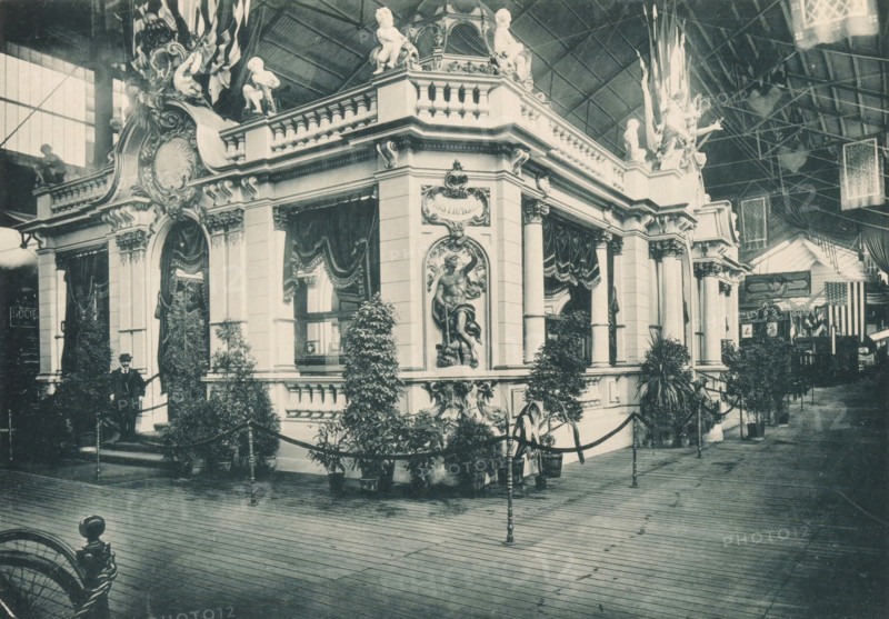 View of the Norddeutscher Lloyd pavilion at the Antwerp World Exhibition in 1894, Pavillon du Norddeutscher Lloyd, Exposition Universelle d'Anvers 1894, page, photomechanical print, photographer: Charles Bernhoeft, Antwerp, cliché maker: Charles Bernhoeft, Luxembourg (Belgian province), 1894, Charles Bernhoeft, photographer, cliché maker, paper, collotype, photoprint, height 153 mm x width 217 mm, palace, world fair, world exhibition, World Exhibition 1894 (Antwerp), Period, 1894-05-05 - 1894-11-05, A large ornate pavilion with classical architectural details occupies the center of the image, featuring columns, balustrades, decorative reliefs, and sculpted figures on its roofline; draped curtains hang over several openings and plants in pots line the exterior, with a rope barrier and stanchions bordering the structure; a man in a dark suit and hat stands near one entrance on a small set of steps; the scene is set inside a spacious exhibition hall with a high arched roof and visible met...