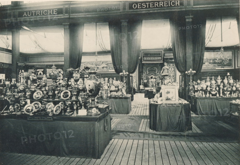 View of the Austrian part of the Antwerp World Exhibition in 1894, Section Autrichienne, Exposition Universelle d'Anvers 1894, page, photomechanical print, photographer: Charles Bernhoeft, Antwerp, cliché maker: Charles Bernhoeft, Luxembourg (Belgian province), 1894, Charles Bernhoeft, photographer, cliché maker, paper, collotype, photoprint, height 153 mm x width 222 mm, Print on third unnumbered page after page 42., counter, shop, container of ceramics: jar, jug, pot, vase, dish, plate, saucer, world fair, world exhibition, World Exhibition 1894 (Antwerp), Period, 1894-05-05 - 1894-11-05, A wide interior scene shows multiple display tables covered with dark cloths holding numerous ornate vases, plates, urns, figurines, and decorative objects arranged closely together, with a large decorative urn placed prominently in the foreground. Tall columns rise toward the ceiling between heavy draped curtains that frame an open central passage leading to another room where a statue stands. Chan...
