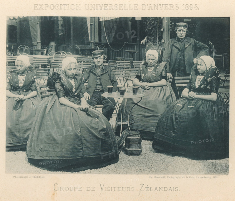 Group of visitors from Zeeland at the Antwerp World Exhibition in 1894, Groupe de Visiteurs Zélandais, Exposition Universelle d'Anvers 1894, page, photomechanical print, photographer: Charles Bernhoeft, Antwerp, cliché maker: Charles Bernhoeft, Luxembourg (Belgian province), 1894, Charles Bernhoeft, photographer, cliché maker, paper, collotype, photoprint, height 154 mm x width 217 mm, anonymous historical persons depicted in a group, in a group-portrait, world fair, world exhibition, World Exhibition 1894 (Antwerp), Period, 1894-05-05 - 1894-11-05, Five women and one man sit and stand around folding chairs and small round tables in an outdoor setting. The women wear full skirts, patterned bodices, and white caps, with layered collars and decorative embroidery on their garments. The man wears a dark jacket and a cap and stands with one hand resting on a chair. On the table are several glasses filled with a dark beverage. Behind them are stacked and arranged folding chairs and parts of ...