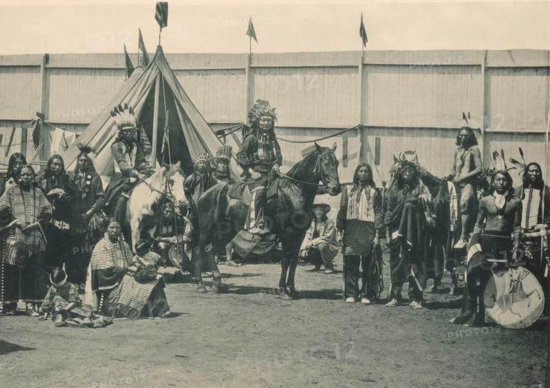 Types Guérriers de la Troupe de Pawnee Bill, Members of Pawnee Bill's group at the Antwerp World Exhibition in 1894, Exposition Universelle d'Anvers 1894, page, photomechanical print, photographer: Charles Bernhoeft, Antwerp, Luxembourg (Belgian province), 1894, paper, collotype, photoprint, height 152 mm x width 219 mm, horse, indigenous peoples of North America, world fair, world exhibition, World Exhibition 1894 (Antwerp), Period, 1894-05-05 - 1894-11-05, A wide group of people arranged in front of a large wooden fence and a canvas tipi, many wearing feathered headdresses and traditional garments, with several individuals mounted on horses and others standing or seated on the ground; visible accessories include beaded necklaces, fringed clothing, patterned blankets, and a round shield resting near one person, while small flags fly atop poles behind the tipi and a variety of long hair styles and face paint appear among the group.