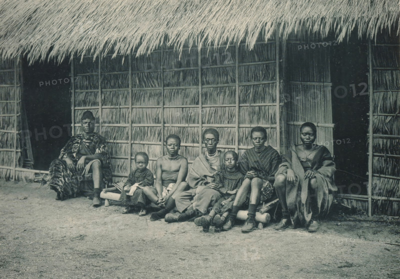 A family sits in front of a house in a recreated Congolese village at the Antwerp World Exhibition in 1894, Famille Congolaise, Exposition Universelle d'Anvers 1894, page, photomechanical print, photographer: Charles Bernhoeft, Antwerp, cliché maker: Charles Bernhoeft, Luxembourg (Belgian province), 1894, Charles Bernhoeft, photographer, cliché maker, paper, collotype, photoprint, height 153 mm x width 216 mm, Print on third unnumbered page after page 74., civic architecture, edifices, dwellings, children (first degree family relationships), world fair, world exhibition, parents (first degree family relationships), World Exhibition 1894 (Antwerp), Period, 1894-05-05 - 1894-11-05, A group of people sit in a row on a low wooden bench in front of a woven thatch wall and a deep overhanging thatch roof, with two dark doorways visible behind them; the thatch panels show vertical and horizontal supports and bundled reeds. The seated figures wear a mix of loose garments and patterned cloth, so...