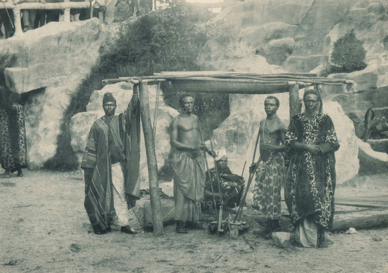 Forgerons du Congo, Five blacksmiths in a recreated Congolese village at the Antwerp World Exhibition in 1894, Exposition Universelle d'Anvers 1894, page, photomechanical print, photographer: Charles Bernhoeft, Antwerp, cliché maker: Charles Bernhoeft, Luxembourg (Belgian province), 1894, Charles Bernhoeft, photographer, cliché maker, paper, collotype, photoprint, height 152 mm x width 216 mm, adult man, smith, blacksmith, world fair, world exhibition, World Exhibition 1894 (Antwerp), Period, 1894-05-05 - 1894-11-05, Five people stand and sit in front of a rocky backdrop beneath a simple wooden frame topped with horizontal poles. One person on the left wears a long striped robe and a pointed hat, holding a vertical post. Next to them a shirtless person stands with a wrapped lower garment and holds a stick. A seated person in the center wears a headwrap and is partially obscured by the structure and a low wooden device with supports. To the right another shirtless person wears patterned...