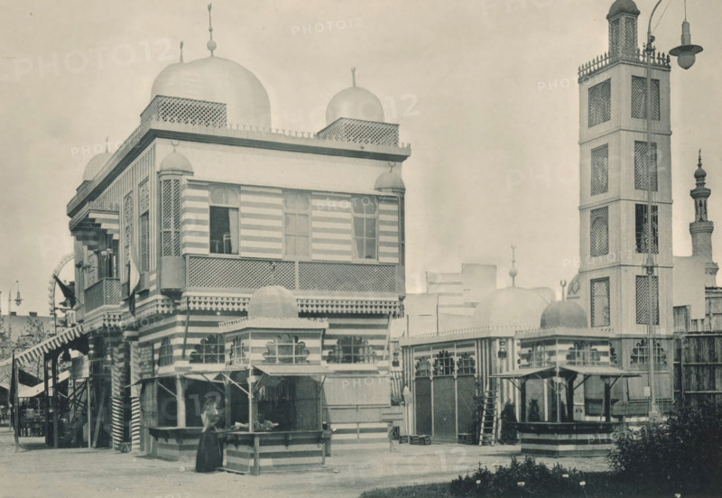 Seccion Marocaine, View of the Moroccan part of the Antwerp World Exhibition in 1894, Exposition Universelle d'Anvers 1894, page, photomechanical print, photographer: Charles Bernhoeft, Antwerp, cliché maker: Charles Bernhoeft, Luxembourg (Belgian province), 1894, Charles Bernhoeft, photographer, cliché maker, paper, collotype, photoprint, height 154 mm x width 222 mm, counter, shop, prospect of village, silhouette of village, world fair, world exhibition, World Exhibition 1894 (Antwerp), Period, 1894-05-05 - 1894-11-05, A sepia-toned street scene showing a multi-story building with striped exterior bands, ornate latticework, rounded domes on the roof, and decorative finials, adjacent to a tall tower with patterned panels and a lamp fixture at its top; the foreground includes a small striped kiosk or stall with a person standing beside it, an awninged walkway with stairs, various windows and balcony railings, a gated entrance with arched openings, and low planted greenery along the fro...