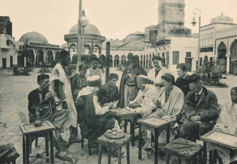 Tunisia, Partie de Cartes devant un Café arabe, Group of men playing a card game on a square in the Tunisian part of the Antwerp World Exhibition in 1894, Exposition Universelle d'Anvers 1894, page, photomechanical print, photographer: Charles Bernhoeft, Antwerp, cliché maker: Charles Bernhoeft, Luxembourg (Belgian province), 1894, Charles Bernhoeft, photographer, cliché maker, paper, collotype, photoprint, height 152 mm x width 217 mm, adult man, card games, square, circus, world fair, world exhibition, World Exhibition 1894 (Antwerp), Period, 1894-05-05 - 1894-11-05, A group of people sit and stand in an open courtyard surrounded by arched buildings and domed structures, with a tall ornate lamp post and a fountain visible in the background; many of the individuals wear headwraps, cloaks, robes, and traditional hats, while one person holds a long curved pipe connected to a vessel on a small table and several others hold small cups; there are low wooden tables and stools with patterned...