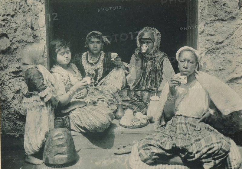 Four women drinking coffee in the Tunisian part of the Antwerp World Exhibition in 1894, Tunisia, Femmes prenant le Café, Exposition Universelle d'Anvers 1894, page, photomechanical print, photographer: Charles Bernhoeft, Antwerp, cliché maker: Charles Bernhoeft, Luxembourg (Belgian province), 1894, Charles Bernhoeft, photographer, cliché maker, paper, collotype, photoprint, height 153 mm x width 216 mm, Print on third unnumbered page after page 90., adult woman, child, cup of coffee, world fair, world exhibition, World Exhibition 1894 (Antwerp), Period, 1894-05-05 - 1894-11-05, Five seated people and a standing child occupy a dark doorway surrounded by rough stone walls, each holding small decorated cups. The figures wear layered, patterned clothing and head coverings in various styles, with jewelry visible on some. A tray with a teapot, lidded container, and stacked cups sits on the ledge in front of them, alongside additional vessels and a large carved head resting on the ground. Th...