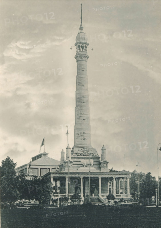 Monument et Exposition de la Compagnie des Produits Kemmerich (Société anonyme) à Anvers, View of the monument to the Compagnie des Produits Kemmerich at the Antwerp World Exhibition in 1894, Exposition Universelle d'Anvers 1894, page, photomechanical print, photographer: Charles Bernhoeft, Antwerp, cliché maker: Charles Bernhoeft, Luxembourg (Belgian province), 1894, Charles Bernhoeft, photographer, cliché maker, paper, collotype, photoprint, height 215 mm x width 152 mm, monument, statue, world fair, world exhibition, World Exhibition 1894 (Antwerp), Period, 1894-05-05 - 1894-11-05, A tall ornate column rises from a multi-tiered stone base topped by a domed turret and slender spire, with decorative bands around the shaft and sculptural elements near the base, set against a cloudy sky; below the column is a classical building with a colonnaded portico, balustrades, and sculpted figures, flanked by flagpoles and trees, with a foreground of lawn and pathways.