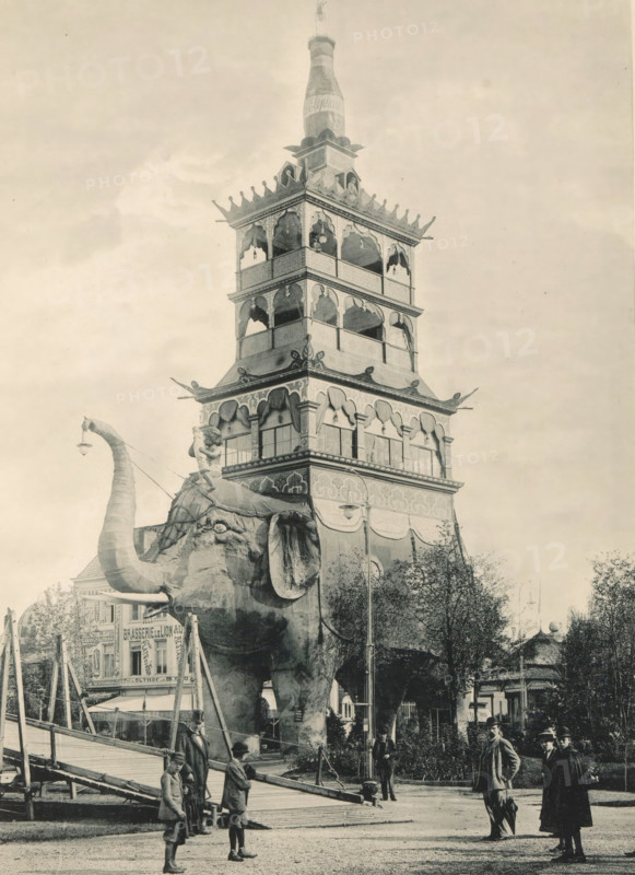 View of an elephant with a building on its back, recreated at the Antwerp World Exhibition in 1894, L'Eléphant Géant, Exposition Universelle d'Anvers 1894, page, photomechanical print, photographer: Charles Bernhoeft, Antwerp, Luxembourg (Belgian province), 1894, paper, collotype, photoprint, height 214 mm x width 153 mm, Print on third unnumbered page after page 94., trunked animals: elephant, civic architecture, edifices, dwellings, world fair, world exhibition, World Exhibition 1894 (Antwerp), Period, 1894-05-05 - 1894-11-05, A large ornate multi-tiered tower rises above a street scene, its tiers decorated with arched openings, carved details, and a bottle-shaped finial at the top; in front of the tower stands a giant elephant figure with a raised trunk and visible tusks, an ornamental howdah on its back, and a small statue mounted atop the howdah; scaffolding and ramps surround the elephant base, while groups of people in period clothing stand and walk nearby on a paved area, some ...