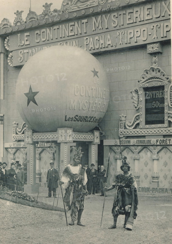 Two men in front of a diorama at the Antwerp World Exhibition in 1894, Le Continent mistérieux, Diorama, Exposition Universelle d'Anvers 1894, page, photomechanical print, photographer: Charles Bernhoeft, Antwerp, cliché maker: Charles Bernhoeft, Luxembourg (Belgian province), 1894, Charles Bernhoeft, photographer, cliché maker, paper, collotype, photoprint, height 215 mm x width 152 mm, adult man, world fair, world exhibition, diorama, World Exhibition 1894 (Antwerp), Period, 1894-05-05 - 1894-11-05, A large spherical object mounted on an ornate platform dominates the upper left, decorated with star shapes and sitting against a façade with carved ornamental trim and patterned panels; two figures in the foreground wear fur and feathered headdresses and hold long staffs while standing on a gravel surface, flanked by formally dressed men and uniformed attendants near the entrance of the decorated building, which features carved columns, scrollwork, and framed openings.