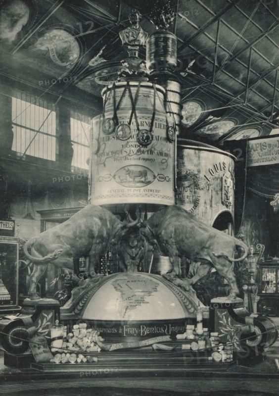 Monument to the Liebig Extract of Meat Company at the Antwerp World Exhibition in 1894, Monument de la Compagnie Liebig, Londres, Exposition Universelle d'Anvers 1894, page, photomechanical print, photographer: Charles Bernhoeft, Antwerp, cliché maker: Charles Bernhoeft, Luxembourg (Belgian province), 1894, Charles Bernhoeft, photographer, cliché maker, paper, collotype, photoprint, height 215 mm x width 152 mm, monument, statue, bull, world fair, world exhibition, World Exhibition 1894 (Antwerp), Period, 1894-05-05 - 1894-11-05, A monochrome indoor scene showing a large decorative display with a domed globe at its base and two life-sized bull sculptures positioned on either side, heads lowered toward the globe. Above the globe a tall cylindrical column rises with ornate embellishments and a bust perched near the top, surrounded by medallions hanging from ribbons. The ceiling overhead has a grid of beams and circular painted panels, and tall windows with vertical panes let light into t...