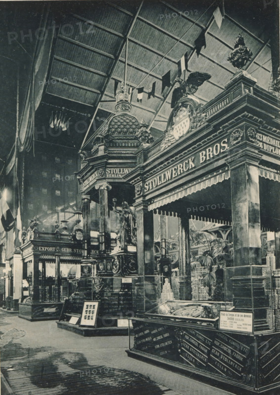 View of a setup of chocolate manufacturer Gebr. Stollwerck from Cologne at the Antwerp World Exhibition in 1894, Pavillons en Chocolat (Gebr. Stollwerck, Koeln.), Exposition Universelle d'Anvers 1894, page, photomechanical print, photographer: Charles Bernhoeft, Antwerp, cliché maker: Charles Bernhoeft, Luxembourg (Belgian province), 1894, Charles Bernhoeft, photographer, cliché maker, paper, collotype, photoprint, height 215 mm x width 152 mm, world fair, world exhibition, chocolate (solid), World Exhibition 1894 (Antwerp), Period, 1894-05-05 - 1894-11-05, A large indoor scene shows an ornate exhibition pavilion with tall columns and decorative capitals supporting an elaborate entablature and dome-like roof element topped with sculptural details; a prominent sculpted eagle sits above a crest set into the building facade and a classical statue stands on a pedestal near the entrance; hanging flags and pennants drape from a high arched ceiling with visible ribs; display cases with glass ...