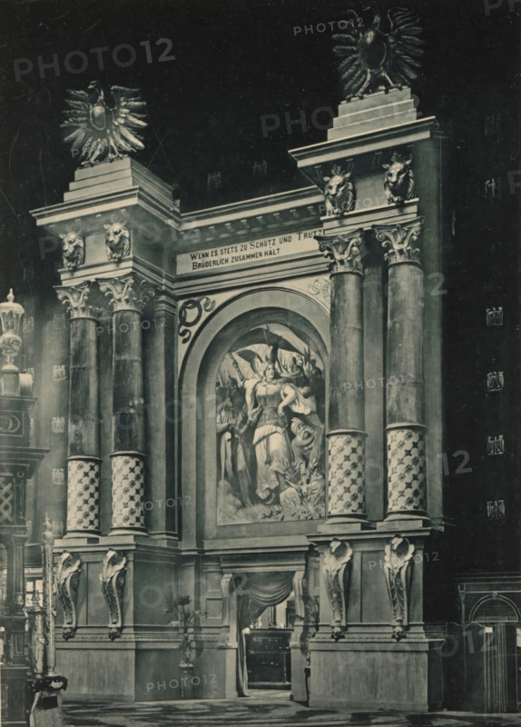 View of the entrance to a music hall in the German part of the Antwerp World Exhibition in 1894, Entrée du Salon de Musique, Section Allemande, Exposition Universelle d'Anvers 1894, page, photomechanical print, photographer: Charles Bernhoeft, Antwerp, cliché maker: Charles Bernhoeft, Luxembourg (Belgian province), 1894, Charles Bernhoeft, photographer, cliché maker, paper, collotype, photoprint, height 215 mm x width 152 mm, gate, entrance, world fair, world exhibition, music-hall, World Exhibition 1894 (Antwerp), Period, 1894-05-05 - 1894-11-05, A grand architectural façade with tall ornate columns and decorative capitals, flanked by sculpted animal heads and topped by large stylized eagles, features a large arched niche containing a painted scene of robed figures and a winged figure; patterned columns with spiral designs rise from a heavy base adorned with carved corbels and draped fabric over a recessed doorway, while ornamental urns, lampposts, and additional sculptural details ar...