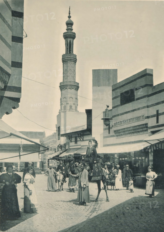 Rue de Caire, View of a street in Cairo, recreated at the Antwerp World Exhibition in 1894, Exposition Universelle d'Anvers 1894, page, photomechanical print, photographer: Charles Bernhoeft, Antwerp, cliché maker: Charles Bernhoeft, Luxembourg (Belgian province), 1894, Charles Bernhoeft, photographer, cliché maker, paper, collotype, photoprint, height 215 mm x width 153 mm, Print on third unnumbered page after page 134., adult man, street, hoofed animals: camel, world fair, world exhibition, World Exhibition 1894 (Antwerp), Period, 1894-05-05 - 1894-11-05, A tall ornate minaret rises behind a street scene, its carved shaft and decorative balconies contrasted against a pale sky, while striped and geometric building facades flank the street with awnings and projecting lamps; in the foreground a group of people stand and walk on a cobbled road, some wearing long robes and others in dresses and jackets, with a man seated on a draped camel and attendants leading other animals, scattered ma...