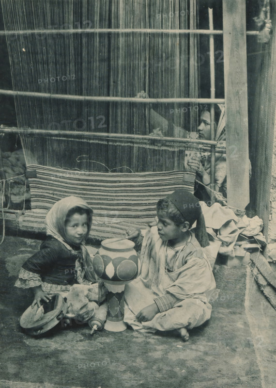 Tunisia, Carpet Manufacturer, Two children playing in front of a woman behind a loom in the Tunisian section of the Antwerp World Exhibition in 1894, Exposition Universelle d'Anvers 1894, page, photomechanical print, photographer: Charles Bernhoeft, Antwerp, clichémaker: Charles Bernhoeft, Luxembourg (Belgian province), 1894, Charles Bernhoeft, photographer, clichémaker, paper, collotype, photogravure, height 215 mm x width 153 mm, tambourine, child, drum (musical instrument), world fair, world exhibition, weaving-loom (women (labourers)), World Exhibition 1894 (Antwerp), Period, 1894-05-05 - 1894-11-05, A woven vertical warp and horizontal rods fill the upper background, with a striped woven textile hanging across the middle; a child peeks through the warp at the right, hand grasping a rod, headscarf visible; in the foreground two children sit on the ground beside a tall painted wooden object, one child wearing a headscarf and layered clothing holding a shallow bowl and a tambourine-l...