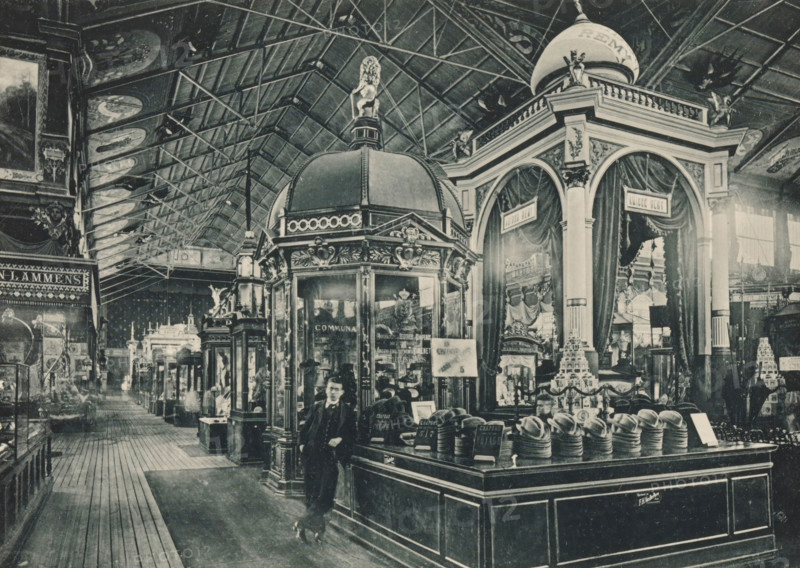 View of the Belgian part of the main exhibition of the Antwerp World Exhibition in 1894, Galerie Centrale, Section belge, Exposition Universelle d'Anvers 1894, page, photomechanical print, photographer: Charles Bernhoeft, Antwerp, cliché maker: Charles Bernhoeft, Luxembourg (Belgian province), 1894, Charles Bernhoeft, photographer, cliché maker, paper, collotype, photoprint, height 153 mm x width 216 mm, Print on third unnumbered page after page 22., counter, shop, world fair, world exhibition, World Exhibition 1894 (Antwerp), Period, 1894-05-05 - 1894-11-05, A large indoor space with a high arched ceiling supported by a visible metal framework, decorative painted panels on the underside of the roof, and a wide wooden plank aisle extending into the distance. In the foreground a tall ornate kiosk with a domed top, sculptural details, glass display windows, and a small sculpted figure atop the dome stands beside a counter piled with stacks of hats arranged on forms. Behind and to the rig...