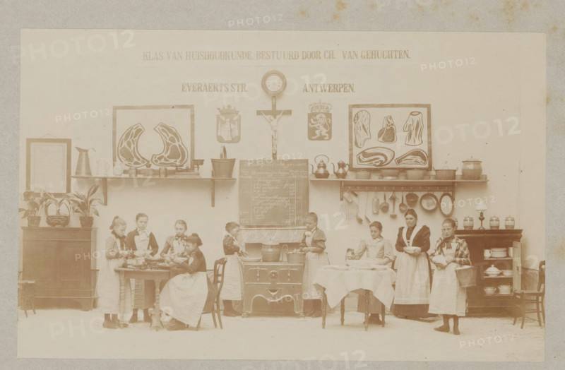 View of a home economics class, photograph, page, Antwerp, c. 1888 - in or before 1893, photographer, paper, daylight collodion silver print, height 106 mm x width 170 mm, cooking food, elementary school, class, form, household chores, A faded sepia photograph of an interior scene showing a wall with framed pictures, a clock, a crucifix, shelves with pots and pans, and hooks holding kitchen utensils; a central stove with a pot, a chalkboard above it, and a table covered with a cloth in front; a group of young girls and a woman wearing aprons standing and seated around tables and the stove, some holding bowls or utensils and some engaged with food on the tables; cabinets and sideboards on either side with jugs, dishes, and potted plants arranged on top; decorative plaques and framed meat diagrams mounted on the wall above the shelves.