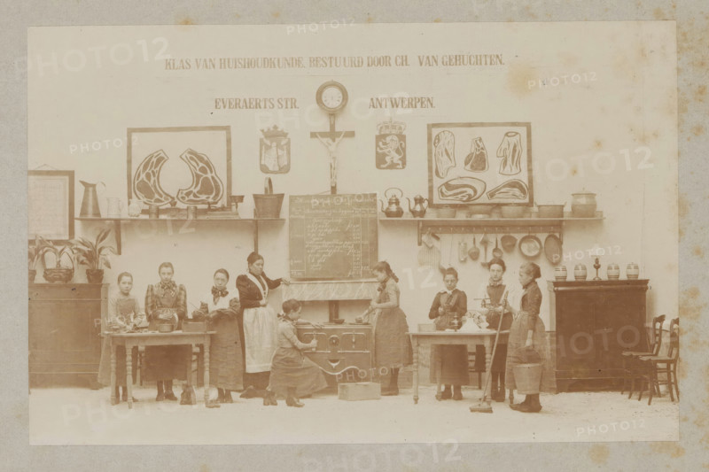 View of a home economics class, photograph, page, Antwerp, c. 1888 - in or before 1893, photographer, paper, daylight collodion silver print, height 105 mm x width 164 mm, elementary school, class, form, household chores, A sepia-toned indoor scene shows a row of young women and girls standing and working along a wall with shelves and framed illustrations, a crucifix, a wall clock, and decorative shields above them. The wall shelves hold metal pots, kettles, pans, and other kitchen utensils, and there are framed anatomical-style prints depicting cross-sections of meat. In front of the wall are tables, a stove, and a small blackboard propped on a mantel; various cooking tools and containers sit on the tables and stove. Several potted plants occupy a cabinet on the left, and jars and a lamp sit atop a cabinet on the right beside stacked chairs. The women wear long dresses and aprons; some hold utensils, a baking pan, a mop, or a bucket, while one kneels at the stove and another stands be...
