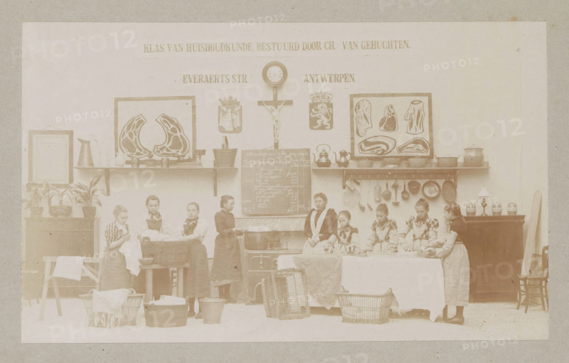 View of a home economics class, photograph, page, Antwerp, c. 1888 - in or before 1893, photographer, paper, daylight collodion silver print, height 104 mm x width 171 mm, elementary school, class, form, laundering, household chores, A faded sepia scene showing a room with a long shelf mounted on the wall holding framed pictures, framed diagrams of cuts of meat, pitchers, pots, kettles, and various kitchen utensils hanging beneath the shelf; a central wall clock above a crucifix and two heraldic shields; a chalkboard with handwritten entries mounted below the shelf; a group of girls standing and seated along the front of the room wearing aprons and dresses, some handling cloths and laundry items, with basins, wooden tubs, baskets, and a washboard on the floor; potted plants and a framed item on a side cabinet at one end and a lamp and jars on a cabinet at the other end, with a chair leaning against the wall.