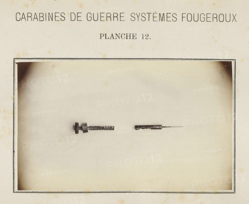 Two parts of the safety catch of a rifle, photograph, page, inscription: 'Carabines de guerres systèmes Fougeroux', number: 'Planche 12.', photographer: Edouard Magis, Liège, c. 1857 - in or before 1867, paper, albumen print, height 158 mm x width 96 mm, Image in section at the back., firearms: rifle, A sepia-toned page with a rectangular bordered area containing two small metal objects centered near the top of the bordered area; the left object is a short cylindrical piece with textured knobs, a coiled spring extending from it, and a small threaded section, while the right object is a slender metal rod attached to a short hollow cylindrical housing with a pointed tip; the page shows slight spotting and aged discoloration around the edges.
