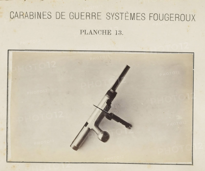 Safety catch of a rifle, photograph, page, inscription: 'Carabines de guerres systèmes Fougeroux', number: 'Planche 13.', photographer: Edouard Magis, Liège, c. 1857 - in or before 1867, paper, albumen print, height 158 mm x width 96 mm, Image in section at the back., firearms: rifle, A rectangular page with a centered light background and a thin double-lined border contains a photograph of a metallic mechanical piece placed diagonally, showing a cylindrical shaft with a rounded knob at one end, a bolt-like threaded tip at the other, and a perpendicular handle with a textured grip and a small flat blade projecting from near its base; the page shows light age spots and slight discoloration.