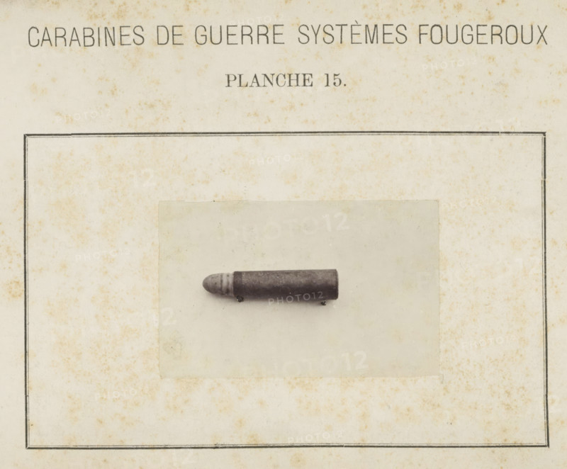 Bullet, photograph, page, inscription: 'Carabines de guerres systèmes Fougeroux', number: 'Planche 15.', photographer: Edouard Magis, Liège, c. 1857 - in or before 1867, paper, albumen print, height 55 mm x width 88 mm, Image in section at the back., ammunition, A rectangular aged paper page with a thin double-lined border and mottled brown spotting, a centered pale rectangular photograph mounted slightly above the middle, and a single dark cylindrical projectile with a rounded grooved tip resting horizontally on the photograph.