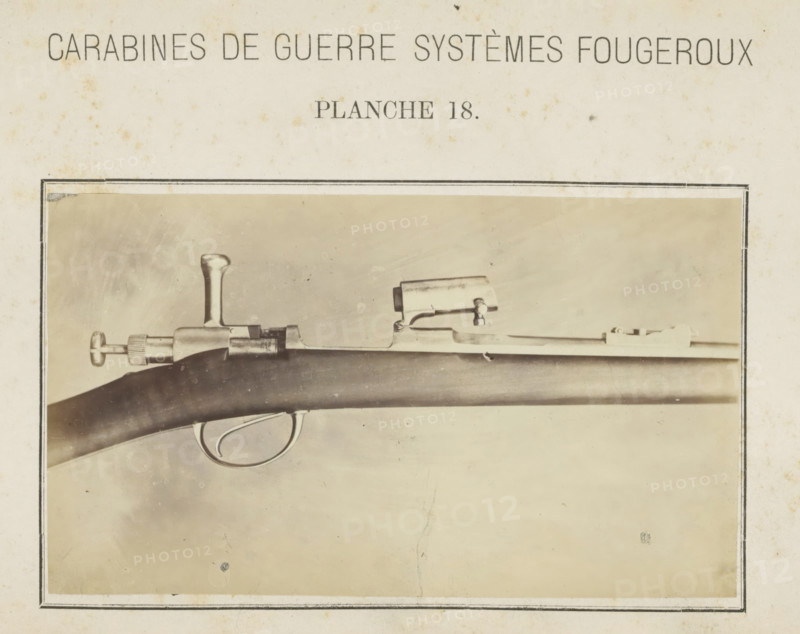 Barrel of a gun, photograph, page, number: 'Planche 18.', inscription: 'Carabines de guerres systèmes Fougeroux', photographer: Edouard Magis, Liège, c. 1857 - in or before 1867, paper, albumen print, height 96 mm x width 158 mm, Image in section at the back., firearms: rifle, A sepia-toned photograph framed by a thin dark border shows a close-up of a firearm mounted horizontally, featuring a dark wooden stock with a smooth curved surface, a metal trigger guard with an oval shape, a metal bolt mechanism with a vertical handle, a cylindrical knurled adjustment knob at the rear, and a rectangular sight mounted atop the barrel; the background is a lighter textured surface with small speckles and faint stains, and the paper surrounding the photograph shows age spots and worn edges.