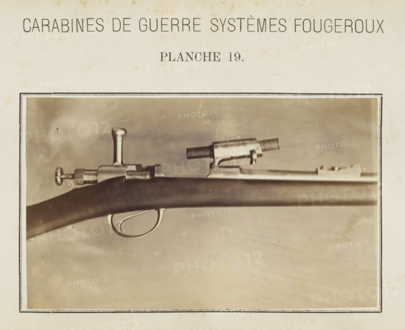 Barrel of a gun, photograph, page, inscription: 'Carabines de guerres systèmes Fougeroux', number: 'Planche 19.', photographer: Edouard Magis, Liège, c. 1857 - in or before 1867, paper, albumen print, height 96 mm x width 158 mm, Image in section at the back., firearms: rifle, A sepia-toned photograph framed by a thin dark border shows a close-up of a rifle horizontal across the image, featuring a wooden stock with a smooth curved trigger guard and visible metal trigger, a bolt mechanism with a rounded handle, a cylindrical sight mounted atop the barrel, and a metallic barrel section extending toward the right; the background is a mottled neutral tone and the surrounding paper shows slight discoloration and spots.