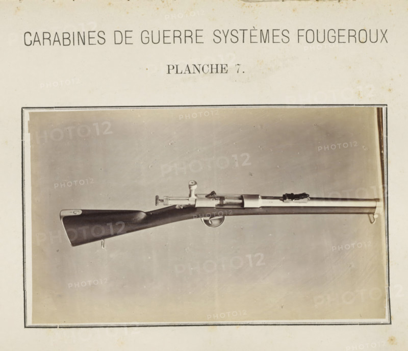 Part of a rifle, photograph, page, inscription: 'Carabines de guerres systèmes Fougeroux', number: 'Planche 7.', photographer: Edouard Magis, Liège, c. 1857 - in or before 1867, paper, albumen print, height 158 mm x width 96 mm, Image in section at the back., firearms: rifle, A black-and-white photograph shows a long rifle mounted horizontally against a plain light background within a rectangular border, featuring a dark wooden stock with a smooth finish and a metal buttplate, a metal trigger guard and curved trigger, a bolt-action mechanism with a round bolt handle, a slender metal barrel with fittings and sighting components, and a sling swivel at the fore end, all centered in the framed image.