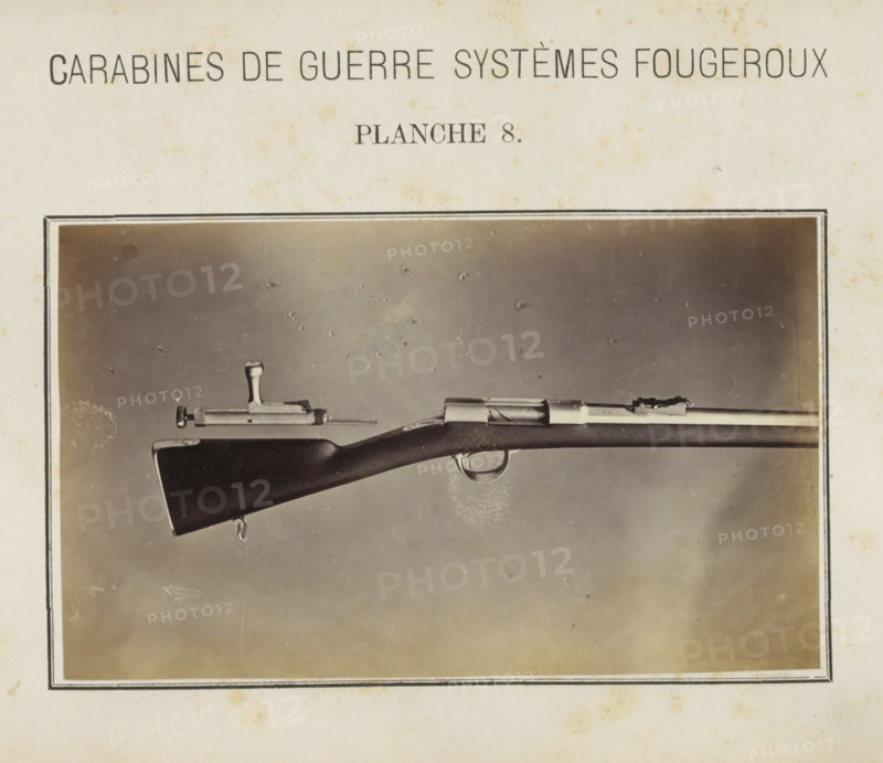 Part of a rifle and a loose safety catch, photograph, page, number: 'Planche 8.', inscription: 'Carabines de guerres systèmes Fougeroux', photographer: Edouard Magis, Liège, c. 1857 - in or before 1867, paper, albumen print, height 158 mm x width 96 mm, Image in section at the back., firearms: rifle, A sepia-toned photograph of a rifle displayed horizontally within a dark rectangular frame, showing the rear stock, trigger guard, and part of the barrel against a mottled background; the wooden stock appears dark and smooth with a metal sling swivel at its end, the bolt mechanism and sights are visible along the top of the barrel, and a detached metal bolt component rests above the rifle; the surrounding paper shows light aging spots and a narrow double-lined border around the photograph.