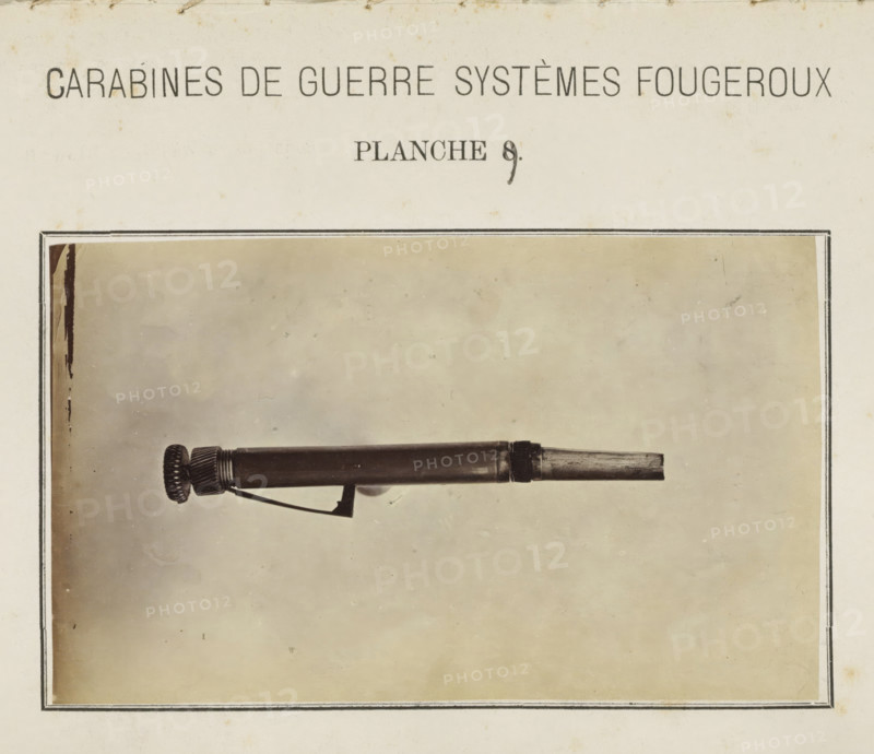 Pal of a rifle, photograph, page, number: 'Planche 9.', inscription: 'Carabines de guerres systèmes Fougeroux', photographer: Edouard Magis, Liège, c. 1857 - in or before 1867, paper, albumen print, height 158 mm x width 96 mm, Image in section at the back., firearms: rifle, A sepia-toned page shows a rectangular framed photograph of a cylindrical mechanical object placed horizontally against a plain background, with one end featuring a ridged circular knob and a short exposed rod at the opposite end; a thin angled metal lever is attached beneath the cylinder and the page around the photograph has a faint aged texture and slight spotting.