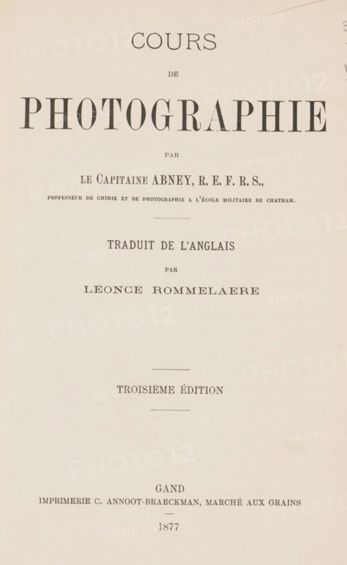 Cours de photographie, par Abney, traduit de l'Anglais par Léonce Rommelaere, book, name: '..Bennert 84.', name: 'L. Bennert, V Bennert', name: 'V Bennert', annotation: 'MAR 1993', writer (clerical worker): Sir William de Wiveleslie Abney, printer (person): C. Annoot-Braeckman, Ghent, 1877, Sir William de Wiveleslie Abney, C. Annoot-Braeckman, writer (clerical worker), printer (person), textile materials, pasteboard, biological material, paper, leather, linen (material), printing, albumen print, spanish marbled paper, stone marbled paper, height 240 mm x width 159 mm x thickness 15 mm, A centered page with a pale beige background showing various lines of dark serif typography arranged vertically, large bold display letters near the top, smaller lines of text spaced below, thin horizontal dividing lines, and faint age spots and slight paper texture across the surface.