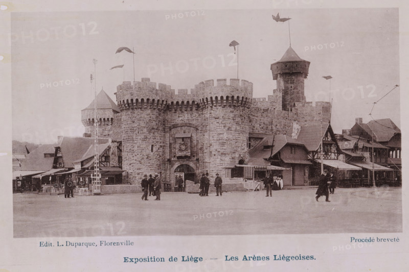 Les Arènes Liègeoises, View of the arena of the 1905 World Exhibition in Liège, Exposition de Liège, photomechanical print, This print is part of a cover with 16 prints., inscription: 'Procédé breveté', Liège, publisher: Louise Duparque, Florenville, 1905, paper, collotype, photoprint, height 98 mm x width 168 mm, world fair, world exhibition, circus ring, World Exhibition 1905 (Liège), A stone fortress facade with rounded battlemented towers and a central arched entrance dominates the scene, flanked by smaller towered and pitched-roof structures. Flags on tall poles and a few decorative pennants extend above the battlements and towers. Timber-framed and awning-covered stalls and buildings line the base of the stone walls, forming a market-like frontage. People in period clothing stand and walk in front of the fortress, including figures near the entrance and pairs or small groups scattered across the open paved area. A tall slender pole with a ladder-like structure stands to one side ...