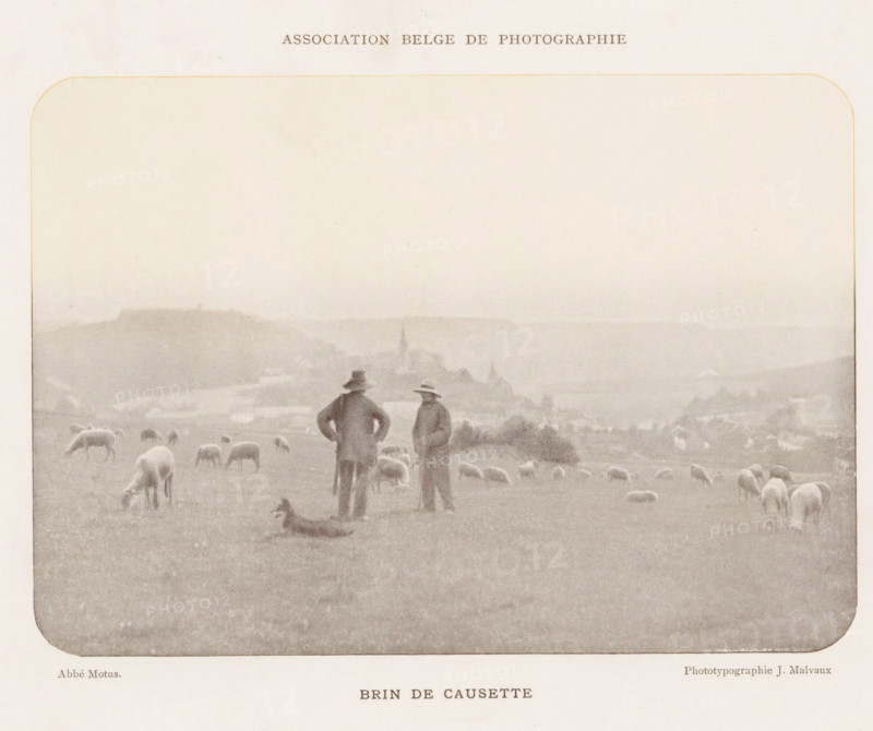 Landscape with sheep herders, Brin de causette, page, photomechanical print, inscription: 'Association Belge de photographie.', photographer: Abbé Motus, cliché maker: Jean Malvaux, Brussels, c. 1893 - in or before 1898, paper, autotypie, height 119 mm x width 168 mm, landscapes, herding, herdsman, herdswoman, shepherd, shepherdess, cowherd, sheep, A sepia-toned rectangular image with rounded corners shows a grassy hillside dotted with grazing sheep and a dog lying on the ground; two men in long coats and hats stand near the center facing slightly toward each other while overlooking a distant village with church spires and low buildings set against rolling hills under a pale sky.