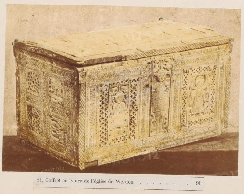 Ivory chest from the St. Luscius Church in Essen-Werden, Coffret en ivoire de l'église de Werden, photograph, page, c. 1877 - in or before 1882, photographer, paper, albumen print, hand coloring, hand colored in yellow, height 111 mm x width 137 mm, chest, coffer, A rectangular carved box with a flat lid sits on a dark surface against a light background, the visible side panels and corner showing intricate repeating geometric and floral patterns with framed rectangular reliefs containing stylized human figures and decorative borders, the top edge of the box featuring a crosshatched band, the surface tones appearing aged and slightly discolored with areas of darker wear along edges and corners.