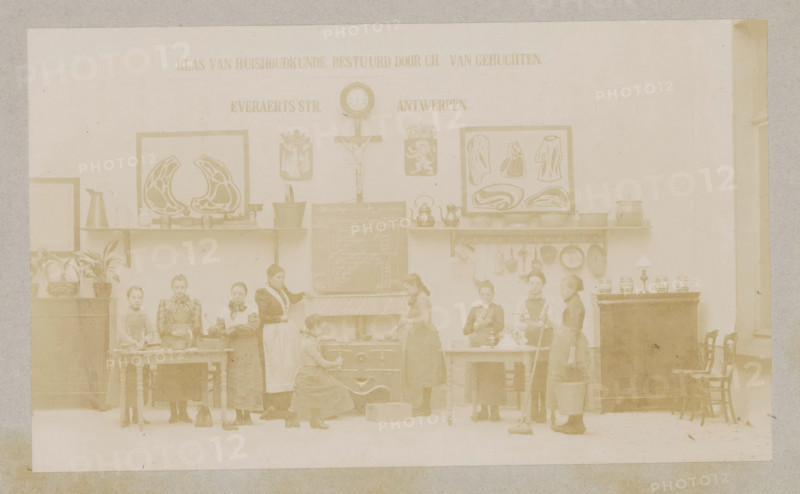 View of a home economics class, photograph, page, Antwerp, c. 1888 - in or before 1893, photographer, paper, daylight collodion silver print, height 104 mm x width 174 mm, elementary school, class, form, household chores, A faded sepia photograph of an interior room showing a long wall with framed decorative pictures, a clock, a crucifix, shelves holding kettles and pots, and hanging pans. Along the lower wall there are tables and a stove with a woman kneeling in front of it, another woman tending the stove, and several girls standing or working at tables holding utensils, bowls, a mop, and a bucket. Potted plants sit on a cabinet at one side and a lamp and jars sit on a tall piece of furniture at the other side, with chairs lined up beside it. The floor is visible and the room has a high window on the right.