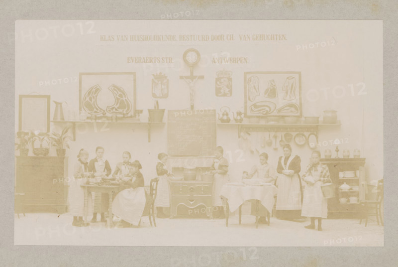 View of a home economics class, photograph, page, Antwerp, c. 1888 - in or before 1893, photographer, paper, daylight collodion silver print, height 104 mm x width 171 mm, cooking food, elementary school, class, form, household chores, A faded sepia photograph showing an interior scene with a long wall as a backdrop, shelves mounted on the wall holding pitchers, pots, kettles, bowls, and framed illustrations; a clock and a crucifix are mounted above a central stove with a chalkboard on top of it; potted plants sit on a cabinet at one side and jars and dishes sit on shelving at the other side; several girls and a woman wearing aprons stand and sit around tables and the stove, some holding bowls, utensils, or a basket, others appearing to be engaged in food preparation; chairs and tables with tablecloths are arranged in front of the stove and along the scene.