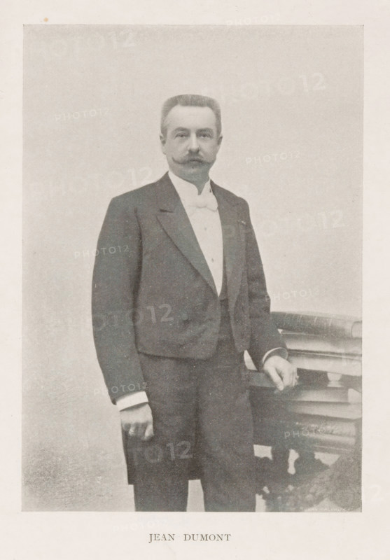 Portrait of Jean Dumont, Jean Dumont, page, photomechanical print, inscription: 'Similigravure.', cliché maker: Jean Malvaux, c. 1899 - in or before 1904, photographer, paper, autotypie, height 145 mm x width 101 mm, Person, historical persons, A sepia-toned portrait shows a man standing next to a small ornate table, wearing a dark suit jacket over a light waistcoat and a high-collared shirt with a bow tie, his hair neatly combed and a styled mustache visible, one hand resting on the table and the other hanging by his side, with a plain background behind him.