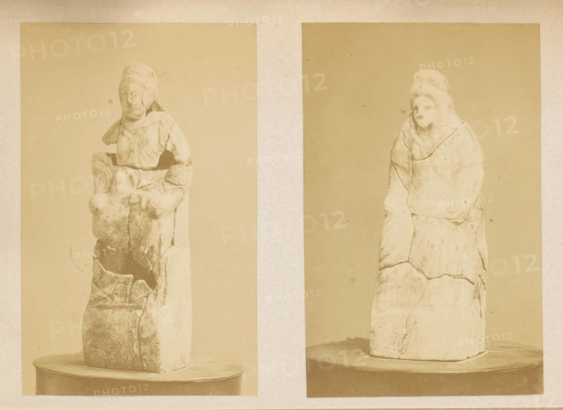 Reconstructed sculpture, excavated at Juslenville in 1868, photograph, page, c. 1868 - in or before 1869, photographer, paper, albumen print, height 116 mm x width 74 mm, Photo on fifth page after page 26., sculpture, Two vertically oriented sepia photographs placed side by side on a textured pale background show tall, weathered stone figures mounted on round bases; the left figure appears fragmented with visible cracks and a worn head with indistinct facial features and a torso showing blocky, broken sections, while the right figure is taller and smoother with a covered head, softened facial features, and layered, cracked surfaces along the body, both images framed by rectangular margins and sharing similar neutral backdrops.