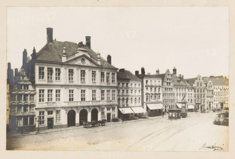 View of the Warehouse on the Korenmarkt in Ghent, photograph, page, annotation: 'Le 'Packhuys' (voir page 17)', photographer: Edmond Sacré, Ghent, c. 1894 - in or before 1899, paper, gelatin silver print, developing gelatin silver print, height 114 mm x width 175 mm, street, large-scale structure for storage, eg: warehouse, depot, silo, A wide street scene showing a row of multi-storey buildings with ornate facades and varying rooflines, some with decorative gables and chimneys, and a large classical building with tall windows and arched ground-floor openings in the foreground, horse-drawn carts parked along the curb, small awnings extending over sidewalk entrances, a freestanding kiosk or pavilion near the street, and an open paved square stretching across the scene under a pale sky.