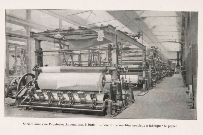 Société anonyme Papeteries Anversoises, à Duffel. - Vue d'une machine continu à fabriquer le papier, View of a paper-making machine at the Société anonyme Papeteries Anversoises in Duffel, page, photomechanical print, Duffel, cliché maker: Jean Malvaux, c. 1906 - in or before 1911, Jean Malvaux, photographer, cliché maker, paper, autotypie, height 100 mm x width 179 mm, Print on second unnumbered page after page 19., paper (writing material), machine driven by electricity, A wide industrial interior with a long row of large mechanical rollers and gears arranged along the floor, connected by metal frames and chains, with sheets of material wound around some rollers and draped fabric hanging above; the space has high ceilings with beams and skylights, tall vertical supports, and a clear aisle running alongside the machinery toward the far end; various crank handles, wheels, and pulleys are visible on the equipment, and the scene is rendered in a monochrome palette.