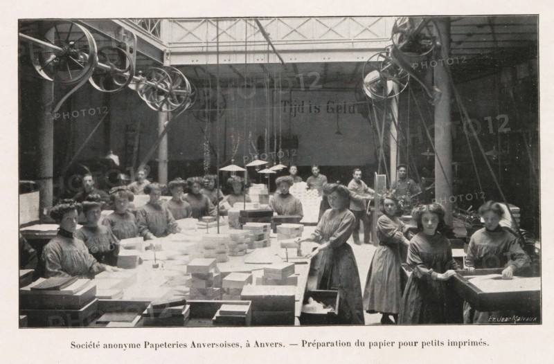 Société anonyme Papeteries Anversoises, à Duffel. - Vue d'une machine continuation à fabriquer le papier, Group of women preparing stacks of paper for small printing in the Société anonyme Papeteries Anversoises in Antwerp, page, photomechanical print, Antwerp, cliché maker: Jean Malvaux, c. 1906 - in or before 1911, Jean Malvaux, photographer, cliché maker, paper, autotypie, height 102 mm x width 172 mm, adult woman, paper (writing material), production of printed matter, book-production, A large indoor workspace with high ceilings and visible pulley wheels and belts near the ceiling, rows of long tables covered with stacks of rectangular sheets and boxes, and a group of women wearing long plain dresses standing and seated around the tables, some handling sheets and boxes and others looking toward the camera, with hanging lamps suspended above the central workspace and structural columns and beams visible in the background.