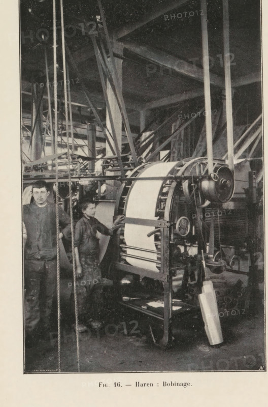 Two men and a paper bending machine in a printing shop in Haren, Belgium, page, photomechanical print, inscription: 'Fig. 16. - Haren: Bobinage.', monogram: 'AN', photographer: unknown, Haren (Brussels), cliché maker: Jean Malvaux, c. 1901 - in or before 1906, Brussels, paper, autotype, height 170 mm x width 110 mm, printing, printing office, adult man, workshop-interior, machine driven by electricity, A tall industrial interior with large beams and support columns fills the background, intersected by a network of ropes, pulleys, and metal frames. In the foreground a large cylindrical machine with a broad wrapped band is mounted on a metal stand with visible gears, wheels, and a drum; a long strip of material hangs from the machine to the floor. Two young boys stand beside the machinery, wearing work clothes and aprons; one boy faces forward holding a rope, the other stands closer to the machine with a hand resting on it, looking toward the first boy. The floor shows scattered tools an...